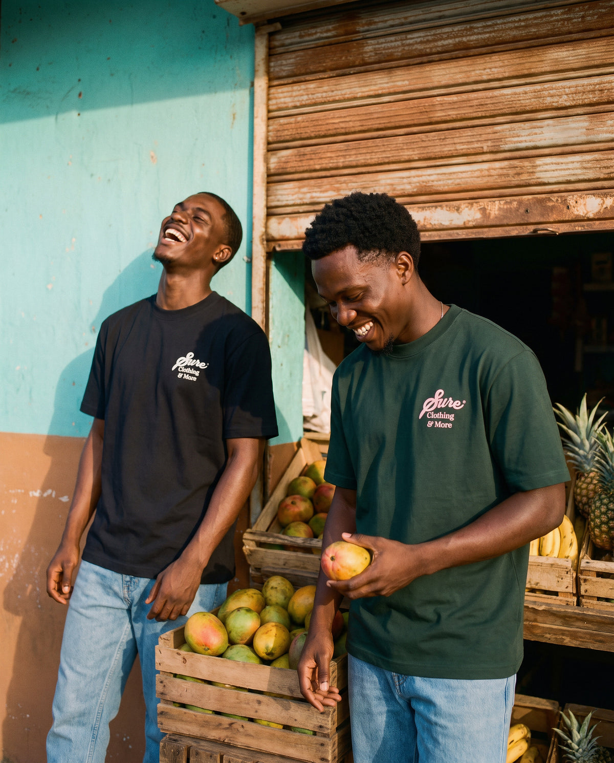 Deux amis qui rient devant les cagettes de fruits d'une épicerie en Guadeloupe, t-shirts Sure noir et vert